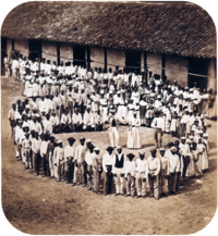 Photograph showing a group of people dressed in white, who have gathered in front of a tile-roofed farm building and observe another large group which has formed a large circle surrounding 5 men straddling large drums, a woman and 2 other men.