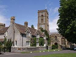 Stone building with a road in the foreground. In the background is the quare tower of a church.