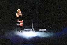 Faraway image of a woman in a short red skilt and black top standing on a stage. Smokes billow around her feet.