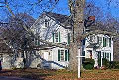 A large white house with green shutters and black roof. In front is a lamppost and large oak tree.