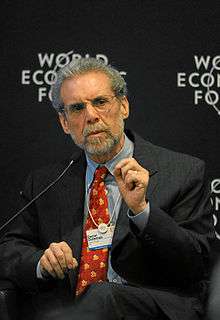 Chest high portrait of man in his sixties wearing a suit, in front of backdrop that says "World Economic Forum"