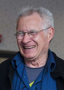 A older man is seen wearing a black coat over a royal blue collared shirt.