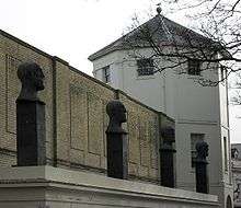 Four evenly spaced sculptures of bald male heads with slightly different facial expressions, each standing on a black rectangular plinth on a flat white surface in front of a yellow-brick wall and an octagonal grey-roofed tower.