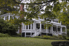 A white wooden house with a roofed front porch seen from slightly downhill, partially obscured by a tree branch