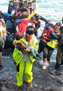 Dr. Alison Thompson is dressed in yellow emergency gear and a love cap. She is carrying a Syrian refugee baby to safety.