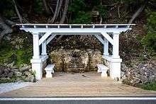 A white, flat-roofed gazebo over a spring, with a plaque located on a rock next to the spring. A short, wide wooden walkway leads from the road to the gazebo.