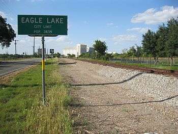 Photo shows the Eagle Lake city limits sign along a road and parallel railroad track. In the distance is a rice dryer.