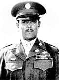 Head and shoulders of a black man, with a carefully trimmed mustache, standing erect and staring directly into the camera. He is wearing a peaked cap and a jacket with three rows of ribbon bars and a pin on the left breast, pins on the lapels, and a braided cord over the left shoulder.