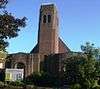 A small brown brick structure unrelieved by dressings or other ornamentation, and partly obscured by shadow, trees and a noticeboard. In the centre is a relatively tall, narrow tower with two arched openings near the top.