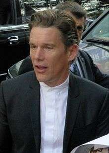 A Caucasian male with dark slick hair, wearing a two-piece gray suit, with a white shirt and black tie.