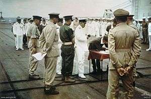 A group of soldiers and sailors parading in various uniforms. In the centre is a small wooden table on which a soldier is signing a document.