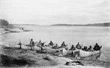 Native people sit in canoes along the shore of a very wide flat river.