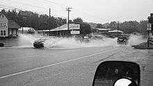 A black-and-white photo of several cars driving in opposite directions on a two-lane road, causing large splashes of water in their wakes