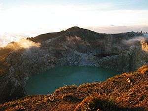 A photograph depicting a blue sky with white clouds at the top, a grey mountain range in the middle, a blue body of water below that, and a rocky terrain in the foreground.