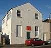 Three-quarter view of a tall, two-storey, plain-walled, white-painted building with a pointed roof. The front has one blank window and a half-blank, half-glazed window flanking a narrow door, above which is an orange sign.  There are also six circular protrusions.  The side wall has three windows and a thin metal flue.