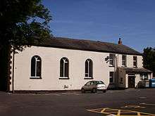 A cream, rendered brick building seen from the south, with dark slate roofs. The right third of the building (looking from the front) is on two storeys, and the remainder is on one.