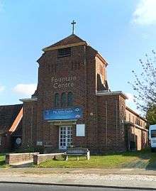 Three-quarter view of a brown-brick church with a short, stubby tower in the foreground.  This is topped with a shallow spire and a crucifix.  There is a concrete bench on a grassed area in front of the building.