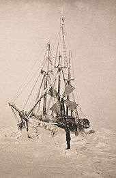 Semi-frontal view of a frost-covered ship surrounded by hummocks of ice. A lone figure stands on the ice nearby.