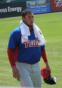 A dark-haired man in a blue baseball jersey with a white towel slung over both shoulders walks on a baseball field carrying a red baseball cap and a black baseball glove together in his left hand.