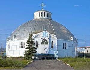 A one-story round building whose walls are white with large blue rectangles topped by a silvery dome with a blue cross and cupola at the center. From the camera a paved walk leads to blue wooden steps going up to its entrance, topped by wooden blue "IHS" letters. A aign in the yard at right says "Our Lady of Victory Church".