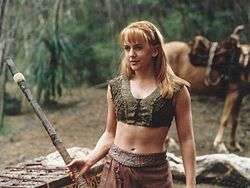 A young blonde woman wearing a green woven tank-top and leather skirt. She holds a wooden fighting staff, a horse can be seen in the background.