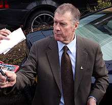 A man in a suit, with a pen in his hand, sigining autographs in front of a vehicle