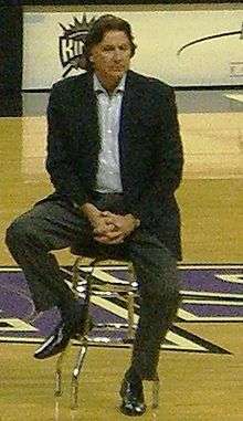 A man wearing a suit sits on a stool at an arena.