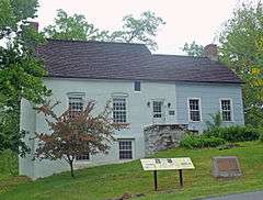 A two-story house in two sections, both with side-gabled shingled roofs, built into a slope towards the right that exposes the basement on the left. The left three bays, including the main entrance with a stone step, is faced in stucco and painted a bluish-grey; the right is wooden and painted light blue. In front there is a memorial plaque on stone and a modern interpretive plaque.