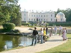 People stand around a small lake, with the back of the palace in the background.