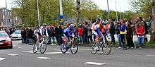 Three road racing cyclists riding in a single file line. Cars follow on the road behind them, and spectators watch from the roadside.