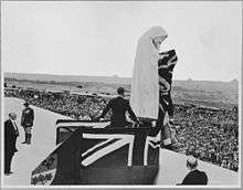 A figure standing on flag covered stage located in from of the statue of Canada Bereft.