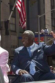 A dark-skinned man in a dark blue pinstriped suit waves with his left hand. A red and white striped flag hangs in the background