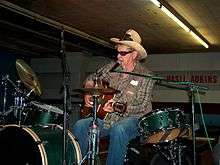 Adkins sitting down, singing, playing guitar with his hands and drums with his feet