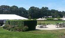 A concrete patio with chairs, tables, and umbrellas, with a hedge and large white tent to the side, and grapevines in the background.