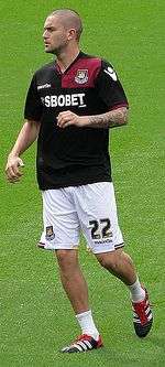 A young man wearing a black and claret shirt, white shorts and white socks, standing on a grass field