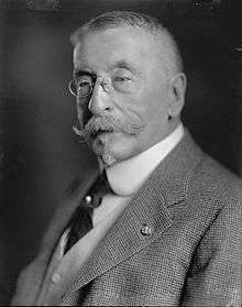 Head and torso of an older white man with a Van Dyke mustache and short hair. He is wearing pince-nez glasses and a checkered suit coat over a vest and dark tie.