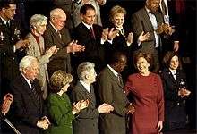 A Native American Hispanic man dressed in a blue suit and red tie stands applauding behind an attractive woman wearing a burgundy-colored dress.