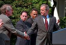 President George W. Bush stands in front of a presidential podium. Standing behind him is a Hispanic-Native American man who is applauding.