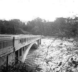 Part of a bridge spans a swollen, fast-moving river. The other part is missing.