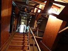 A wooden stairway surrounded by a complicated wooden structural system, rising under a curved wooden ceiling to a distant lit area, illuminated by a bare light bulb