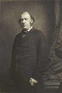 A black-and-white photograph of James Fitzjames Stephen, a balding man with sideburns.  He stands looking to the left of the camera near a curtain.  He is holding a cane in his left hand.