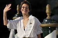 Woman with short, dark hair in a white blouse, smiling and waving