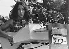 Baez stands behind a too-tall podium bristling with microphones, wearing a plaid sleeveless top, longish hair in a feather cut