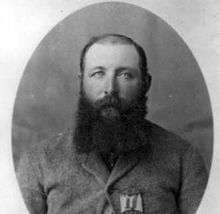 A black and white head and shoulders photograph of a balding white man with a full, dark, beard and a heavy wool coat