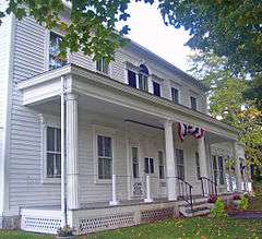 A white house with porch and fluted columns decorated in red, white and blue bunting viewed from its left with some tree branches visible at the sides. A small sign near the front steps says "John Kane House".