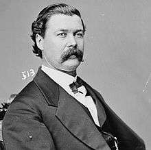 A man with black, wavy hair and a mustache wearing a black jacket and tie and white shirt