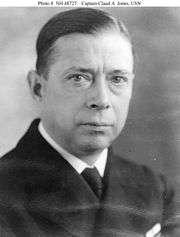 Head of a white man with hair parted and combed back, wearing a dark jacket over a white shirt and dark tie.