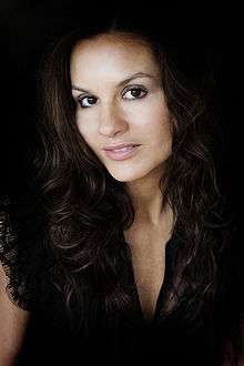 A woman with long brown hair wearing a black shirt looks to the camera