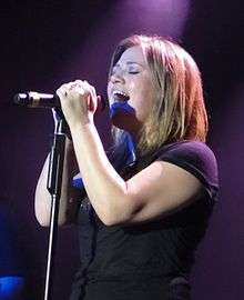 An image of an blonde haired woman, wearing a black T-shirt, singing against a dark violet background