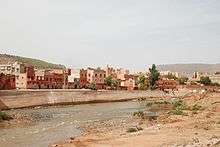 A river runs across the image from left to right with a town in the background, behind a concrete flood defence.  The foreground shows a stony, sparsely vegetated river bank.